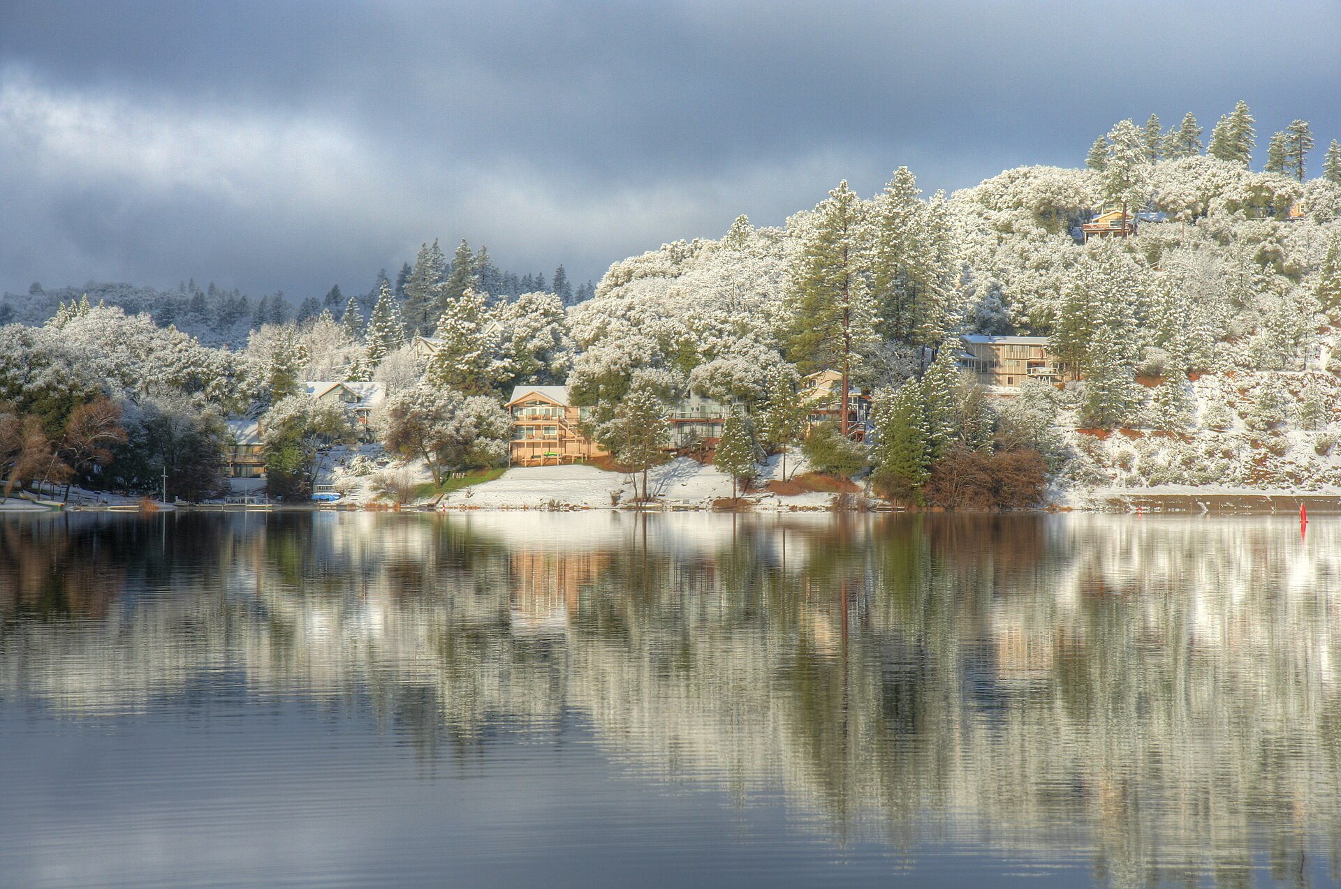 A view across a calm Pine Mountain Lake (Groveland, CA) toward the Dunn Court area. The trees and homes have a light dusting of snow. — California