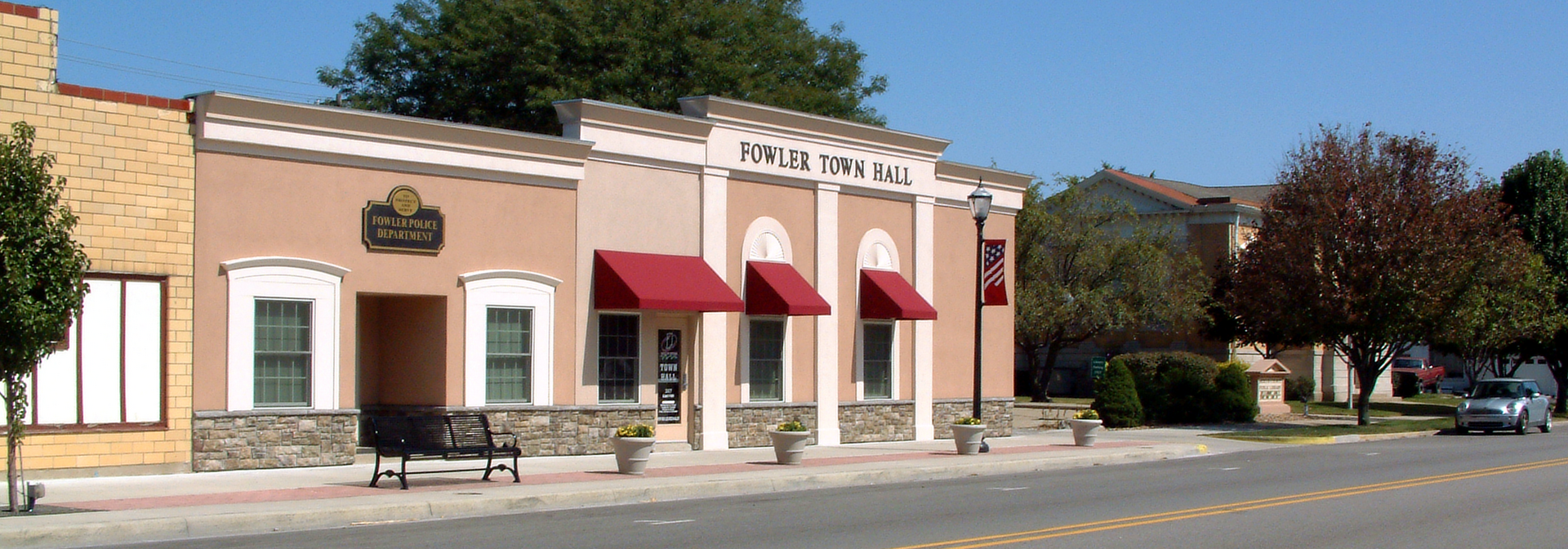 A photo taken on Fifth Street in Fowler, Indiana, looking northeast toward the police department building and the town hall. At right, partly obscured by trees, is the public library.