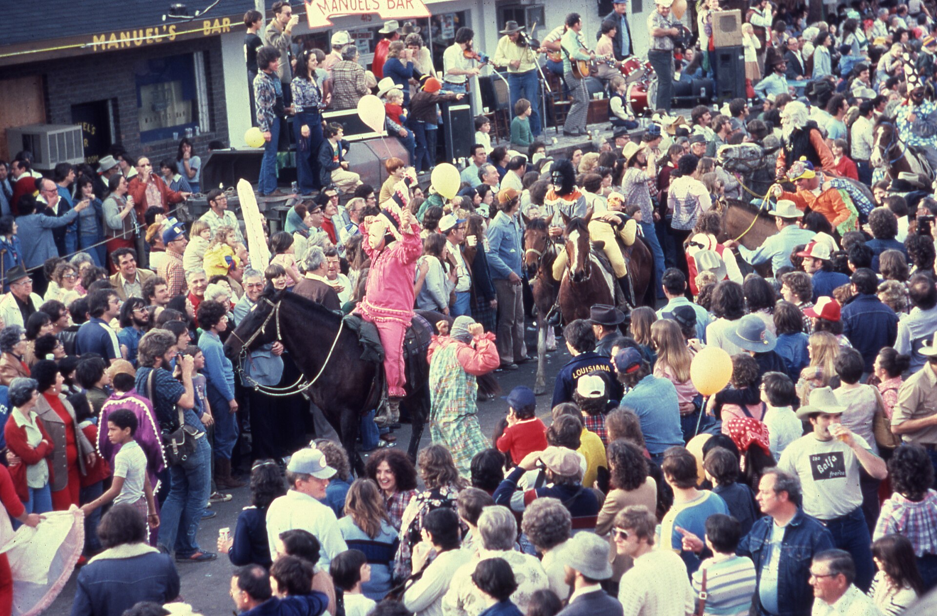 "Parade at Cajun Mardi Gras festival" 1989 Notes: Shows revelers, some riding horses, in traditional Cajun Mardi Gras outfits passing through crowds in front of Manuel's Bar, 6th Street, Mamou, Louisiana.