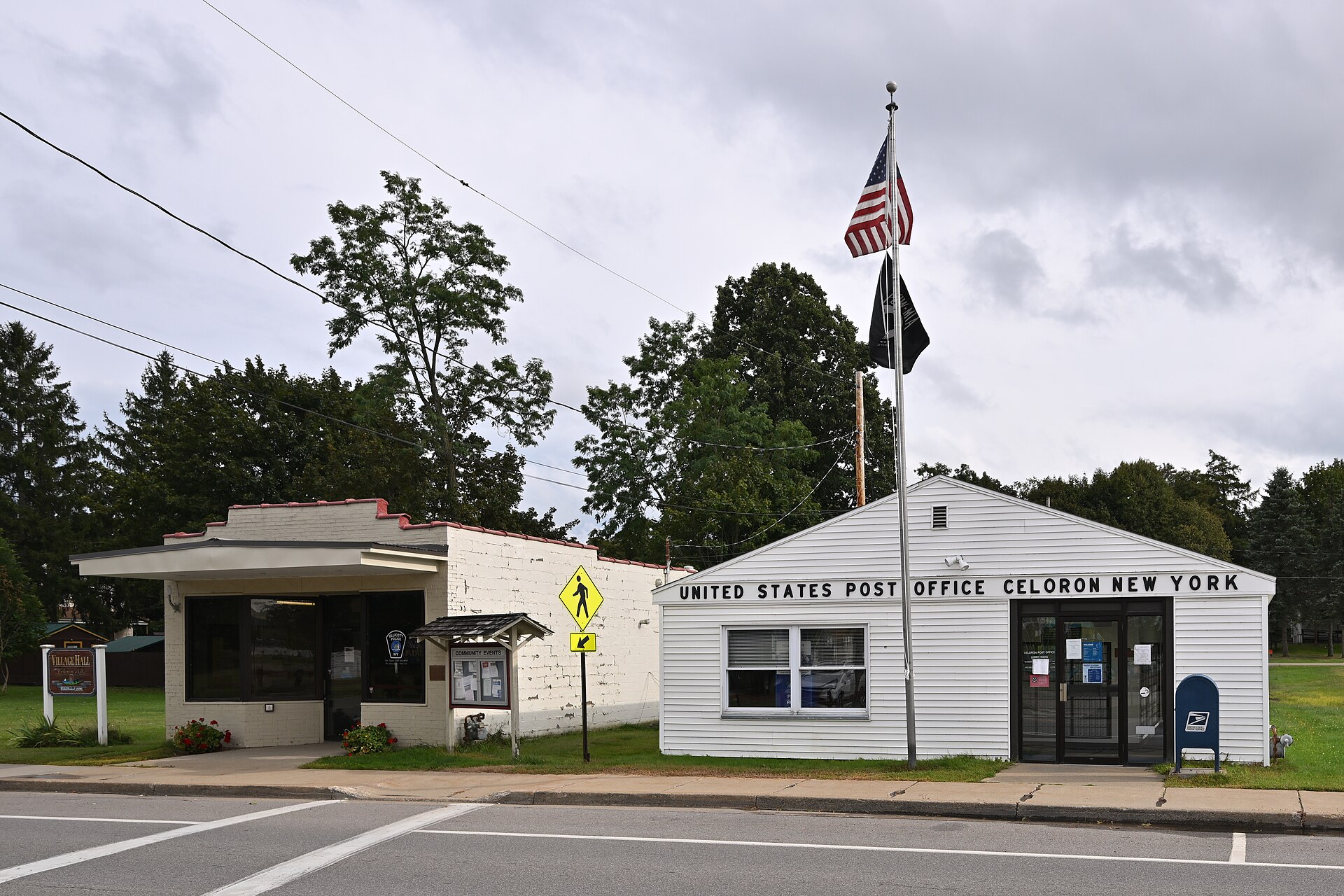A small, white, United States Post Office building with a flag flying in front. To the left is the village hall. 17 Boulevard Avenue, Celoron, New York 14720.